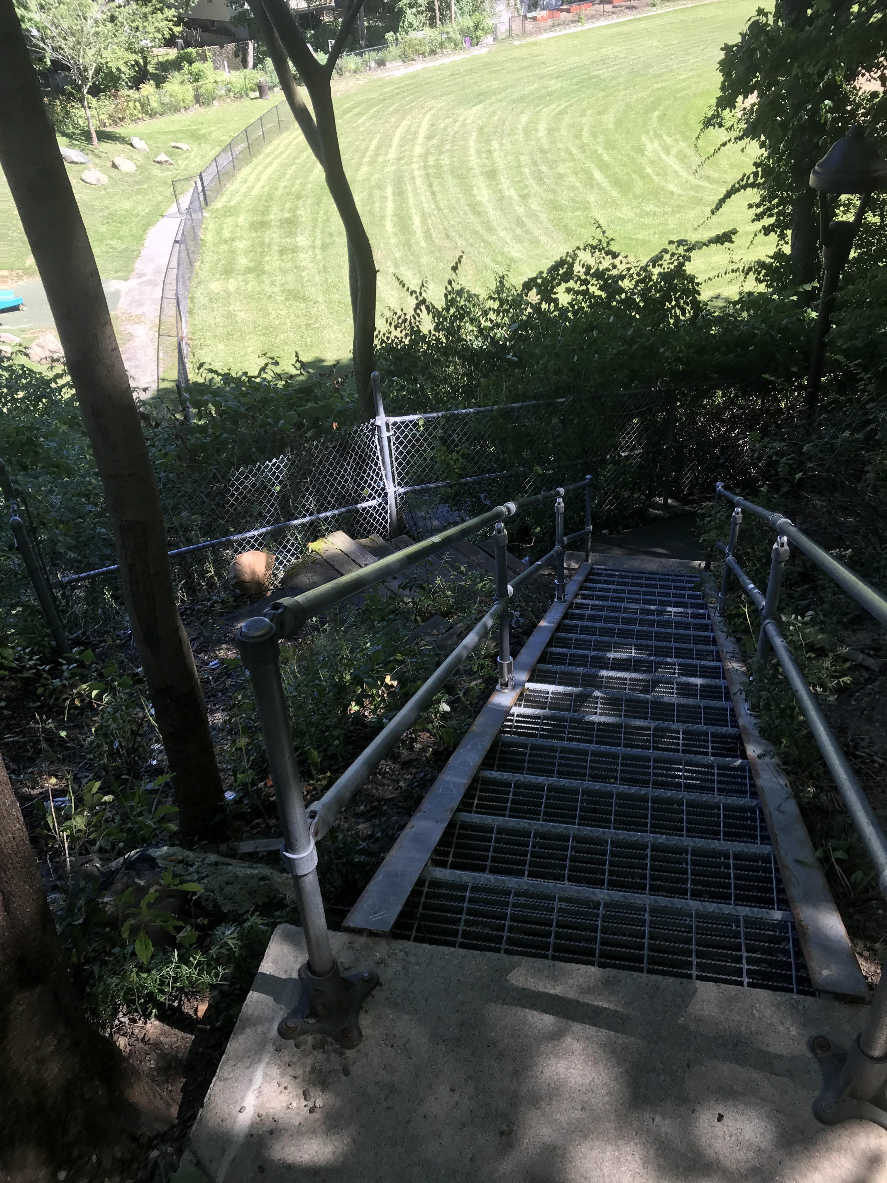 A narrow, steep galvanized-steel stairway with iron pipe railings descends from a concrete platform to the barely-visible edge of a cracked sidewalk below.