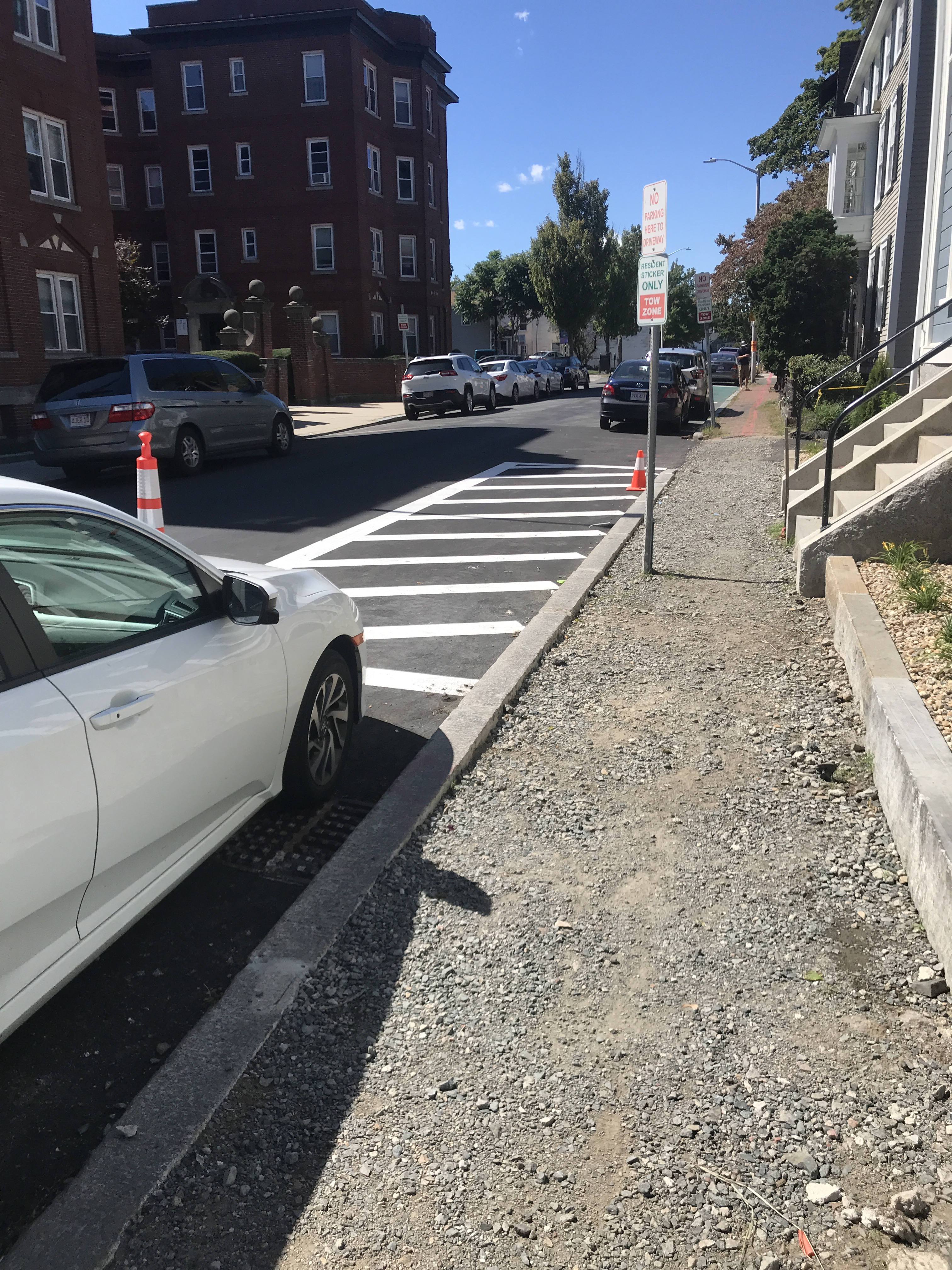 A section of sidewalk has been torn up, exposing a gravel bed. Two caution cones are in the background